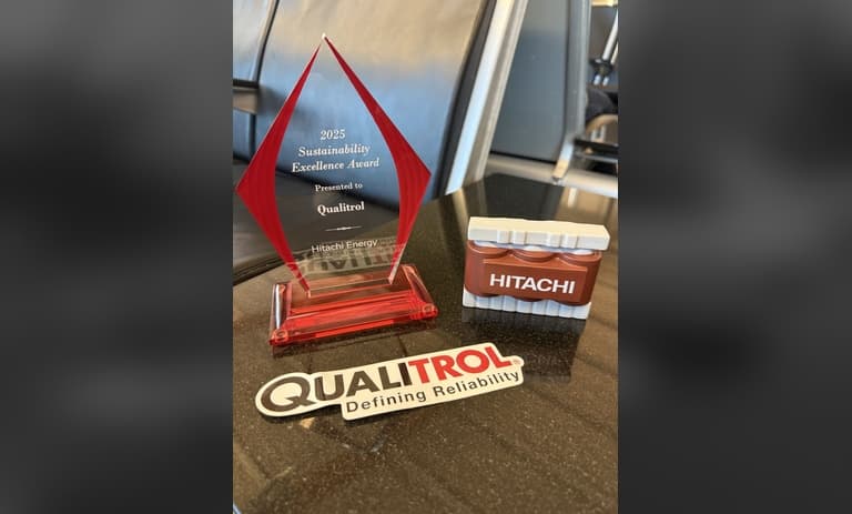 Qualitrol 2025 Sustainability Excellence Award trophy beside a small brown HITACHI transformer model and a Qualitrol ‘Defining Reliability’ sticker on a dark tabletop