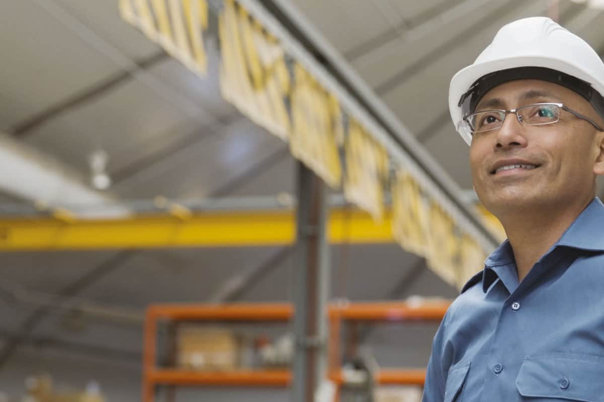 Male engineer in white safety helmet and blue work shirt standing in manufacturing facility with industrial equipment in background